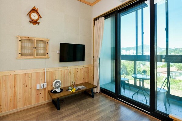 A room with a wooden paneled wall, a flat-screen TV, a wooden bench, a fan, and a large sliding glass door leading to an outdoor balcony with chairs and a table.
