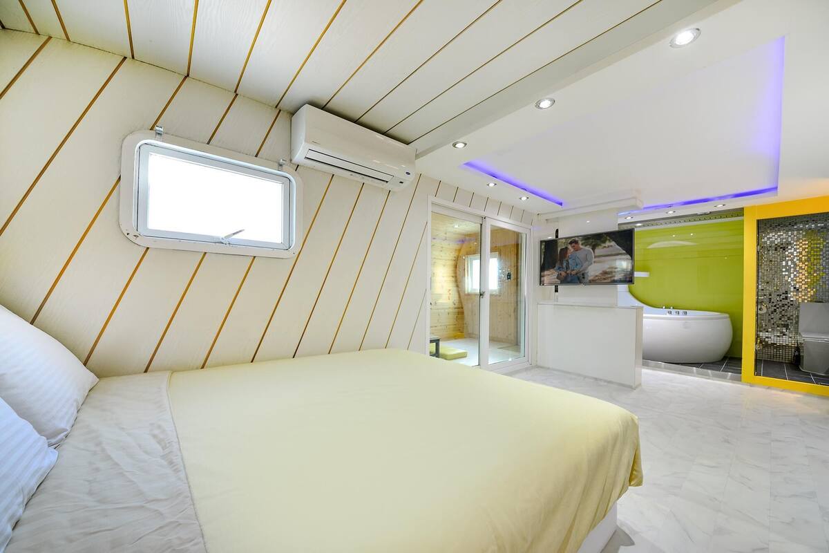 A modern hotel room with a large bed, a small window, an air conditioning unit, a television, and a bathroom with a bathtub.