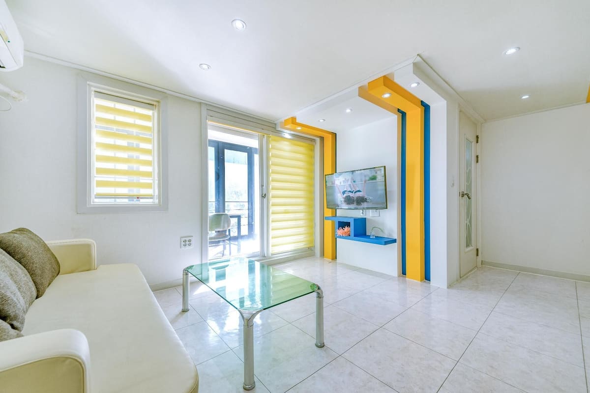 A modern living room with a glass coffee table, a sofa, and a TV mounted on a wall with a yellow accent.