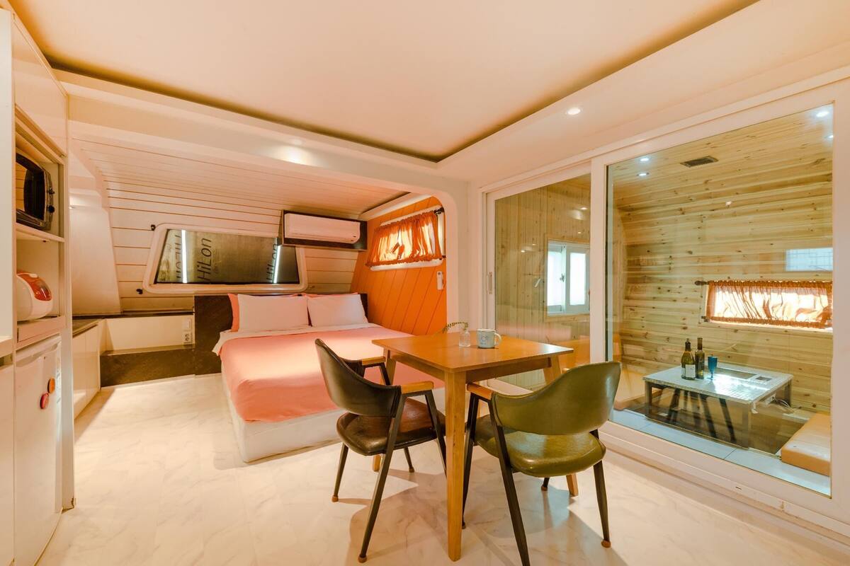 A compact hotel room with a bed, a small kitchenette, a dining area with a table and chairs, and a large window with a view.
