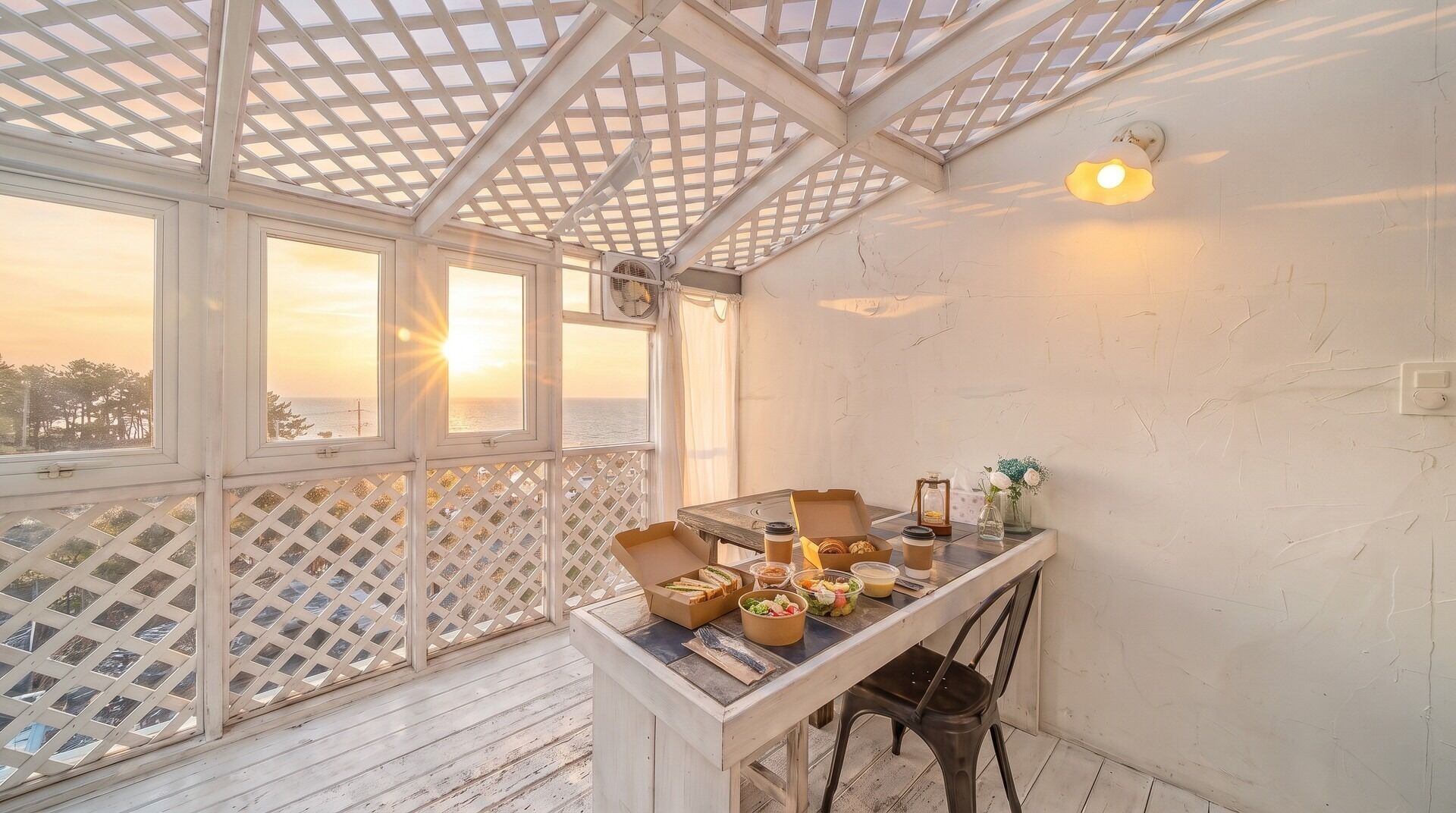 A sunlit room with a wooden table set for breakfast, a view of the sea through large windows, and a white lattice ceiling.