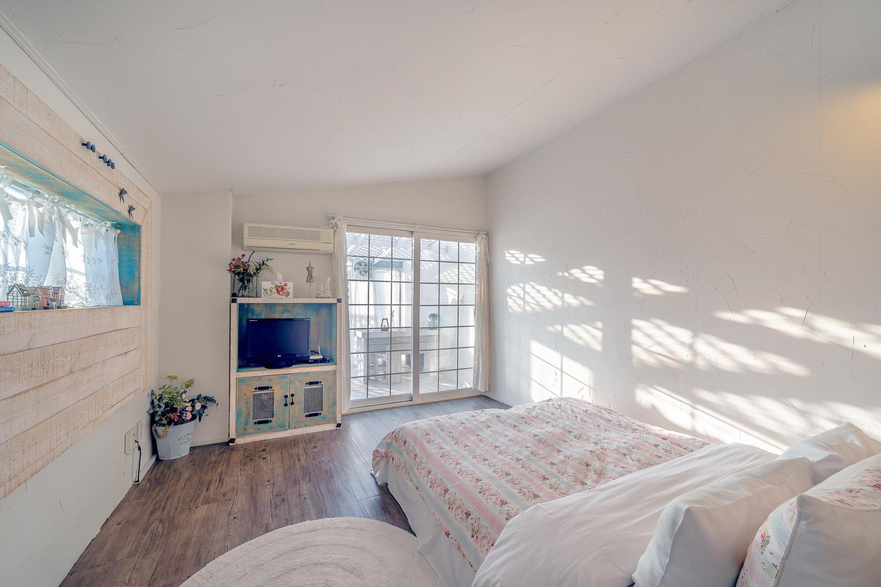A bedroom with a wooden floor, a bed with floral bedding, a TV on a cabinet, and a large window with a view of the outside.