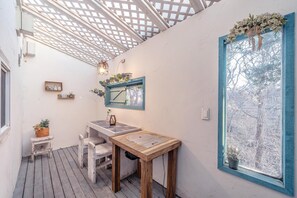 Un espace extérieur accueillant, aménagé avec une table et des chaises en bois, un petit tabouret et une fenêtre donnant sur les arbres.