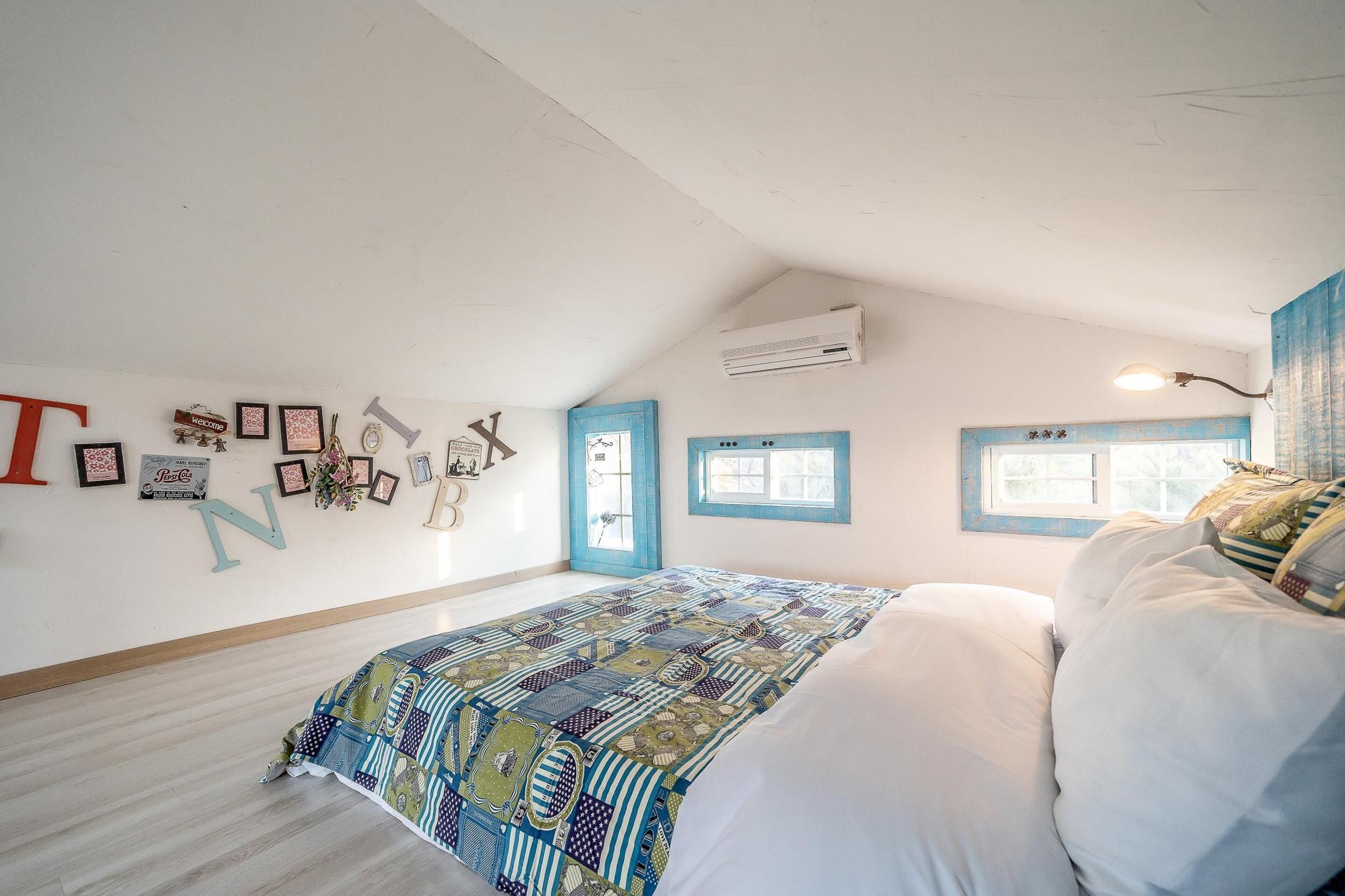 A bedroom with a large bed, a window with blue shutters, and a wall with framed pictures and letters.