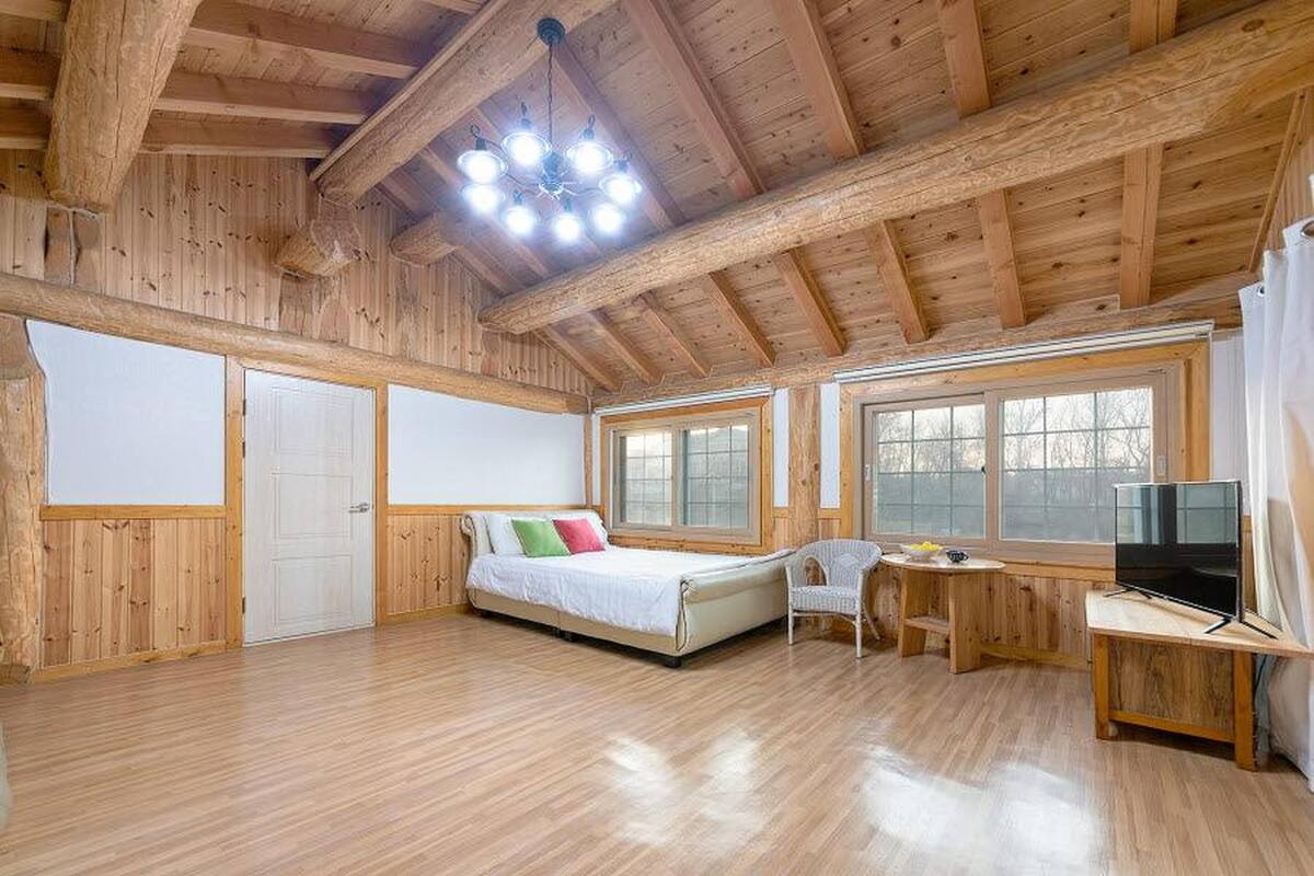 A spacious room with a wooden ceiling, a bed with pillows, a desk with a chair, and a TV.