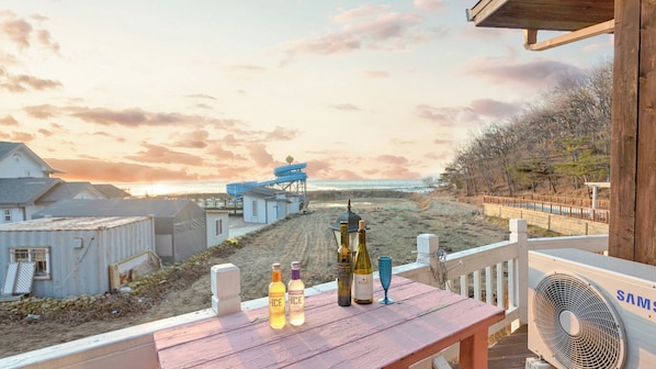 Um deck de madeira com vista para uma praia e oceano, um ventilador e garrafas de bebidas alcoólicas.