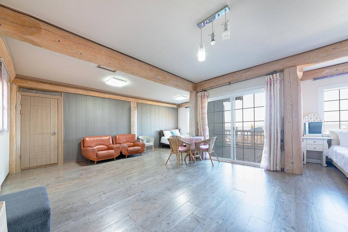 A spacious room with a wooden ceiling, a dining area with a table and chairs, a sofa, a bed, and a large window with curtains.
