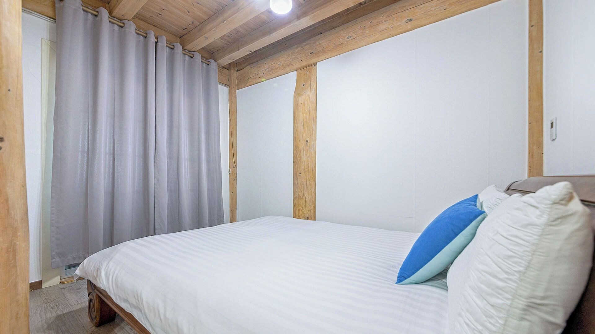 A bedroom with a wooden ceiling, a bed with white bedding and two pillows, and a window with curtains.