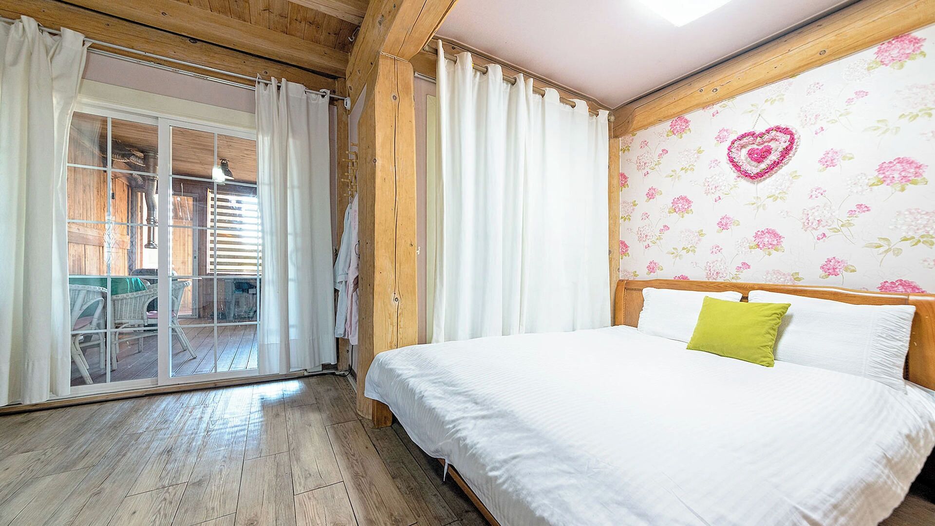 Un dormitorio con piso de madera, una cama con ropa blanca, un almohadón verde y un papel tapiz floral.