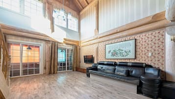 A spacious room with a wooden floor, a leather sofa, a television, and a large window with curtains.