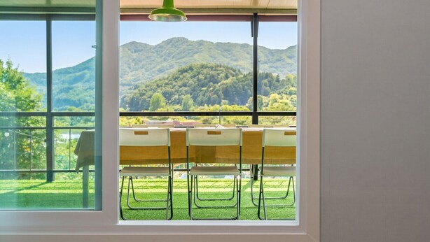 A room with a large window overlooking a mountainous landscape, a table with chairs, and a hanging light fixture. - Gapyeong Four Season Pension (Gapyeong)