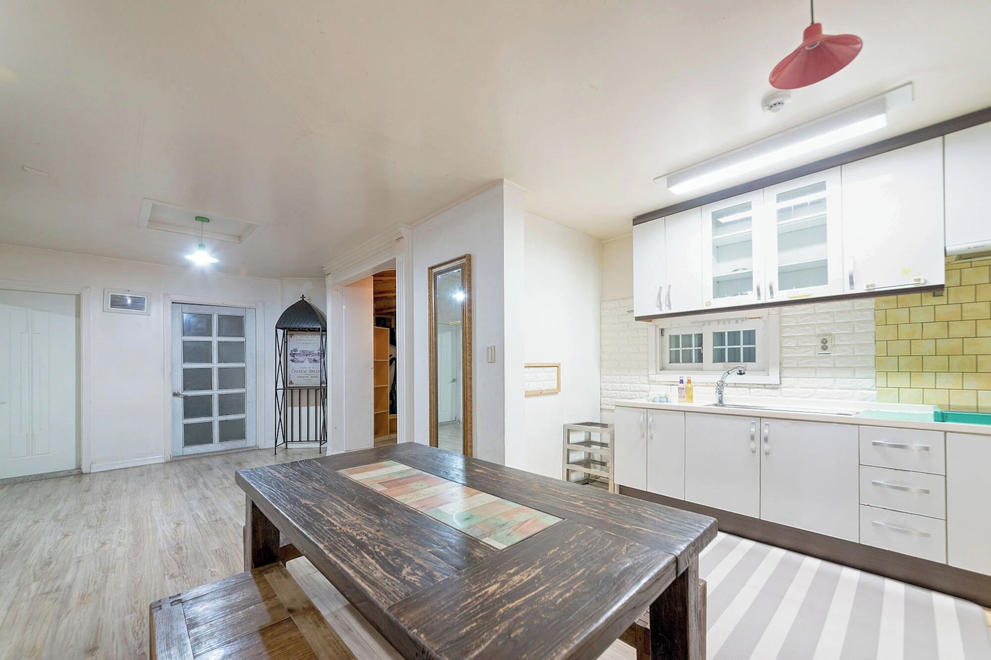Quarto espaçoso com mesa de jantar de madeira, cozinha com armários brancos e uma luminária pendente vermelha.