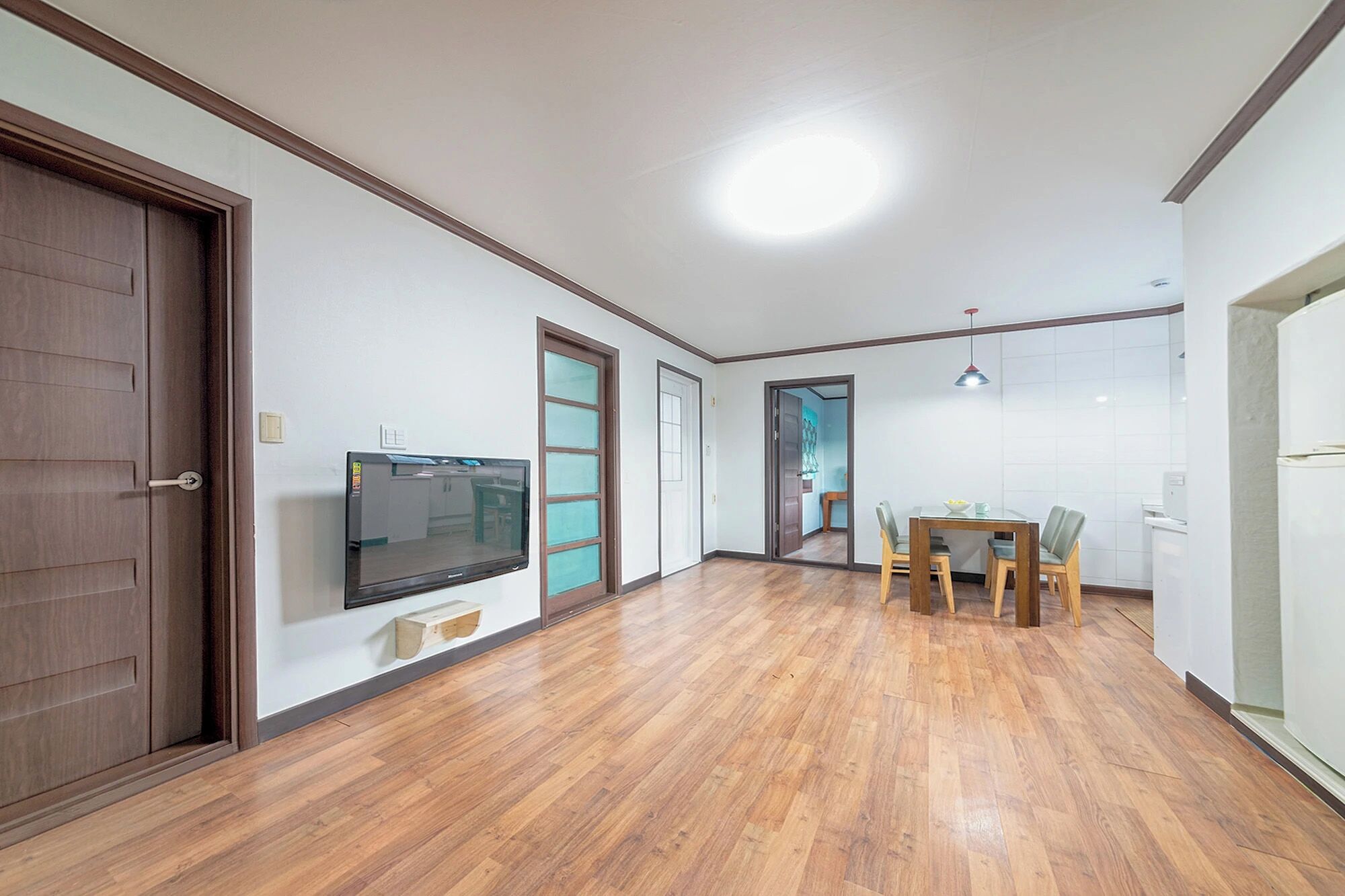 A spacious room with a wooden floor, a dining table with chairs, a television mounted on the wall, and a door leading to another room.