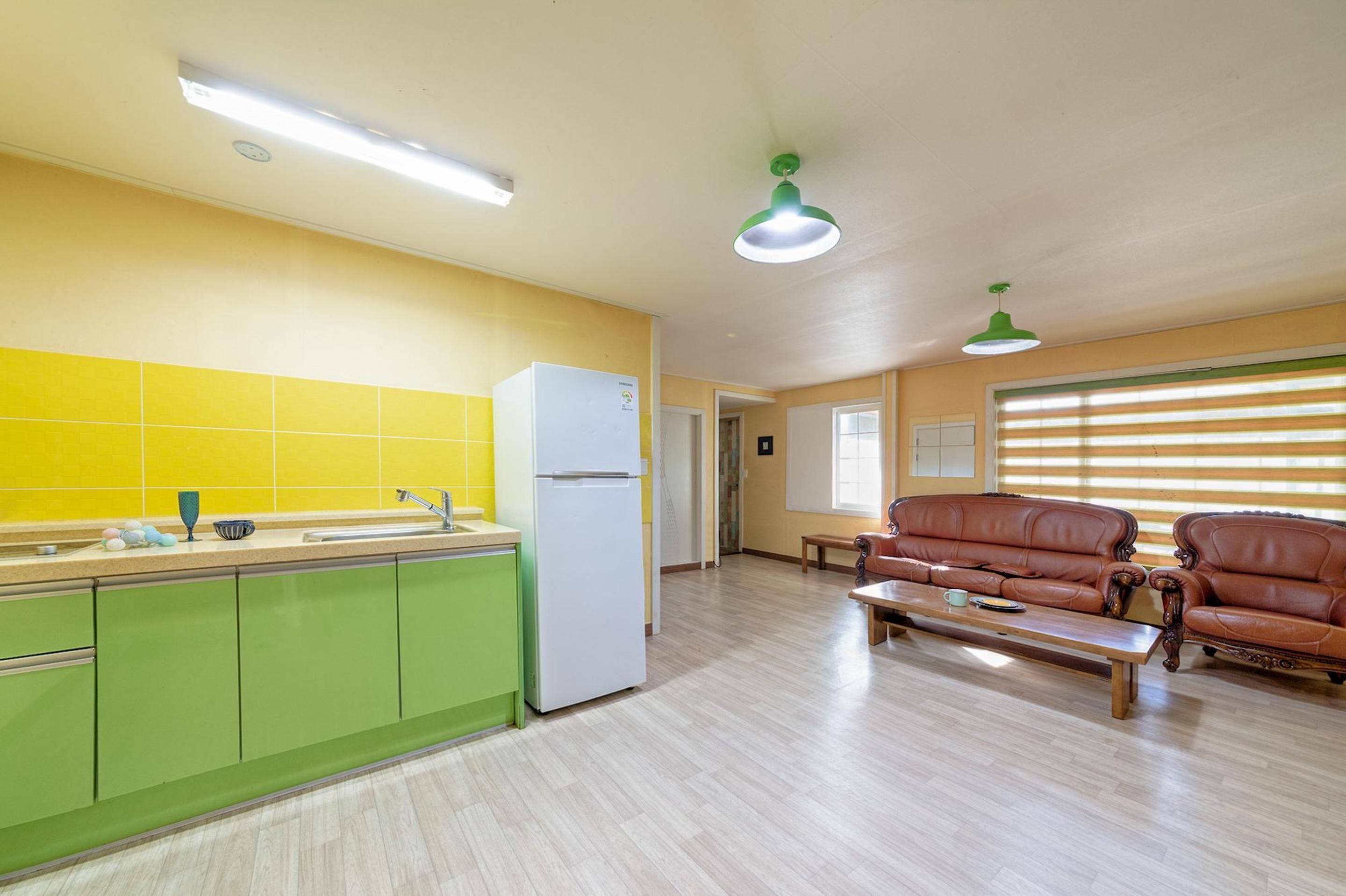 A kitchen with green cabinets, a white refrigerator, and a yellow tiled backsplash. There is a brown leather sofa and a wooden table in the living area.