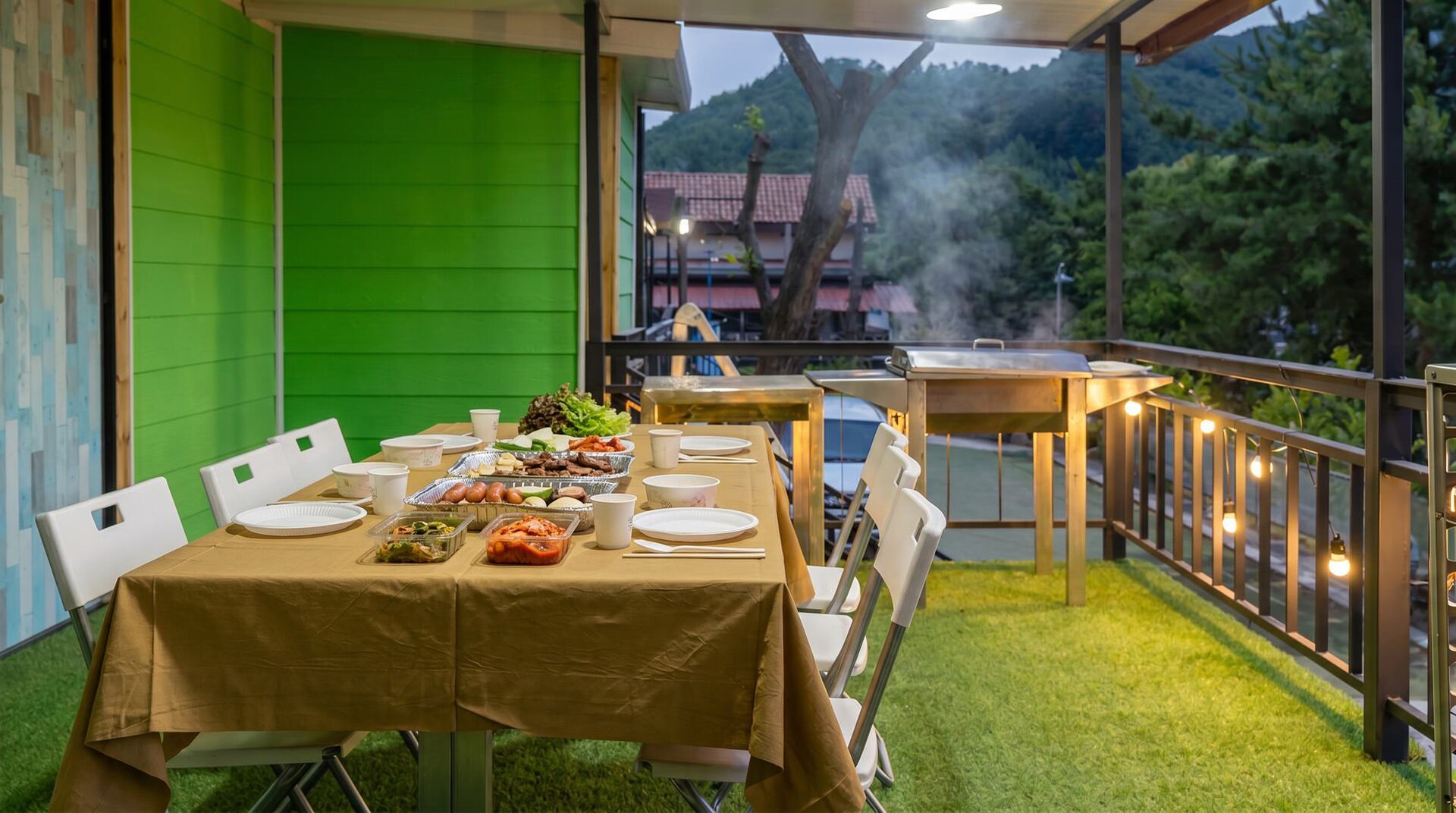 An outdoor dining area with tables set for a meal, surrounded by a green wall and a view of a mountainous landscape.