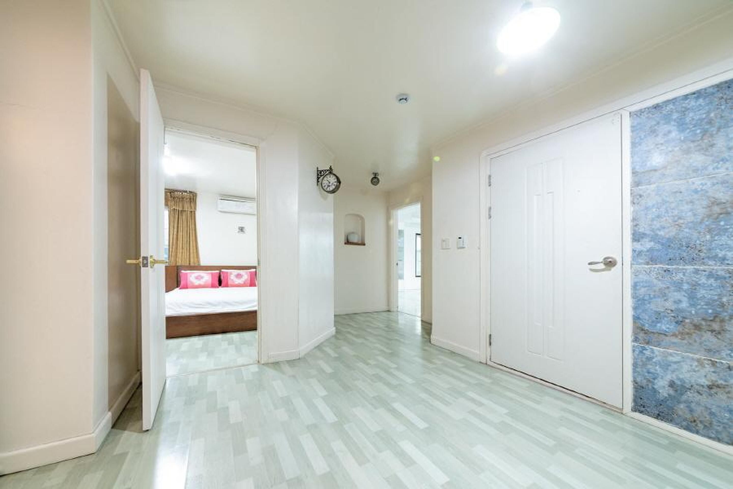 A hallway with a bed in the background, a clock on the wall, and a door with a blue-tiled wall.
