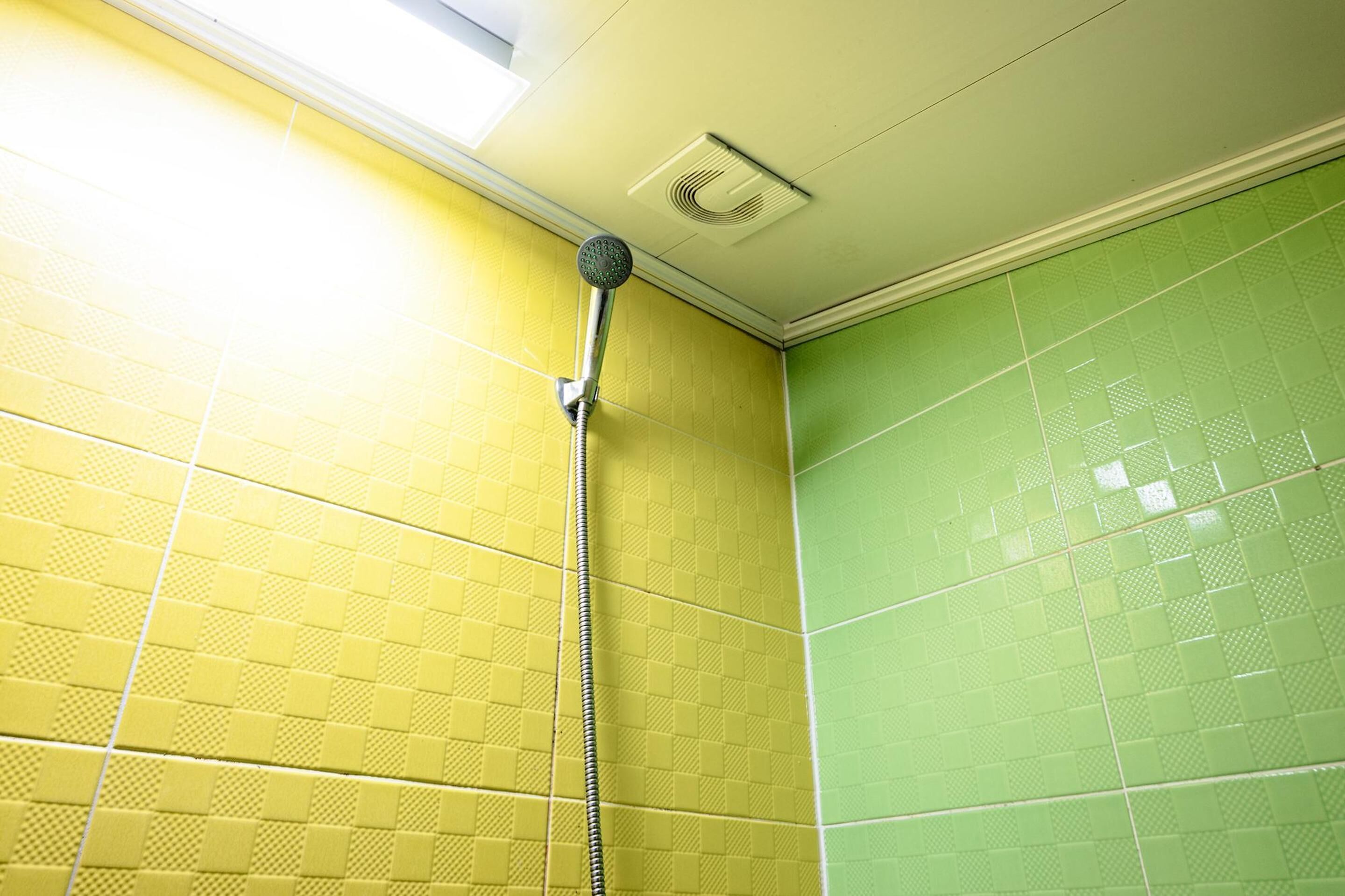A bathroom with yellow and green tiled walls, a showerhead, and a ceiling vent.