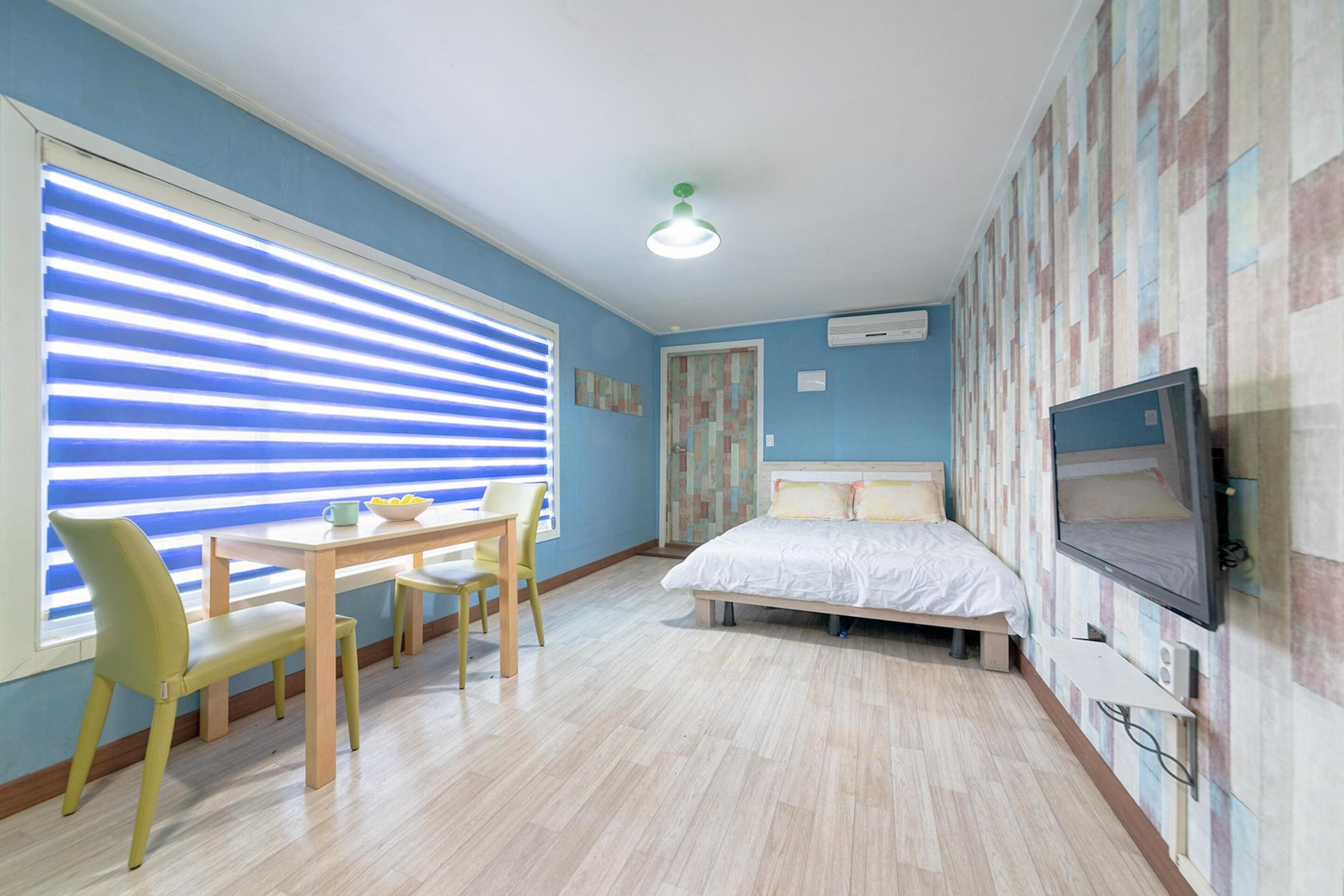 Une chambre à coucher comprenant un lit, une télévision, une fenêtre avec des stores, une table avec des chaises et un ventilateur de plafond.