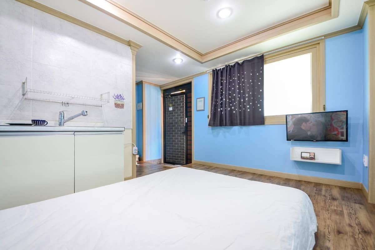 A bedroom with a bed, a TV, a window with curtains, a door, and a kitchenette.
