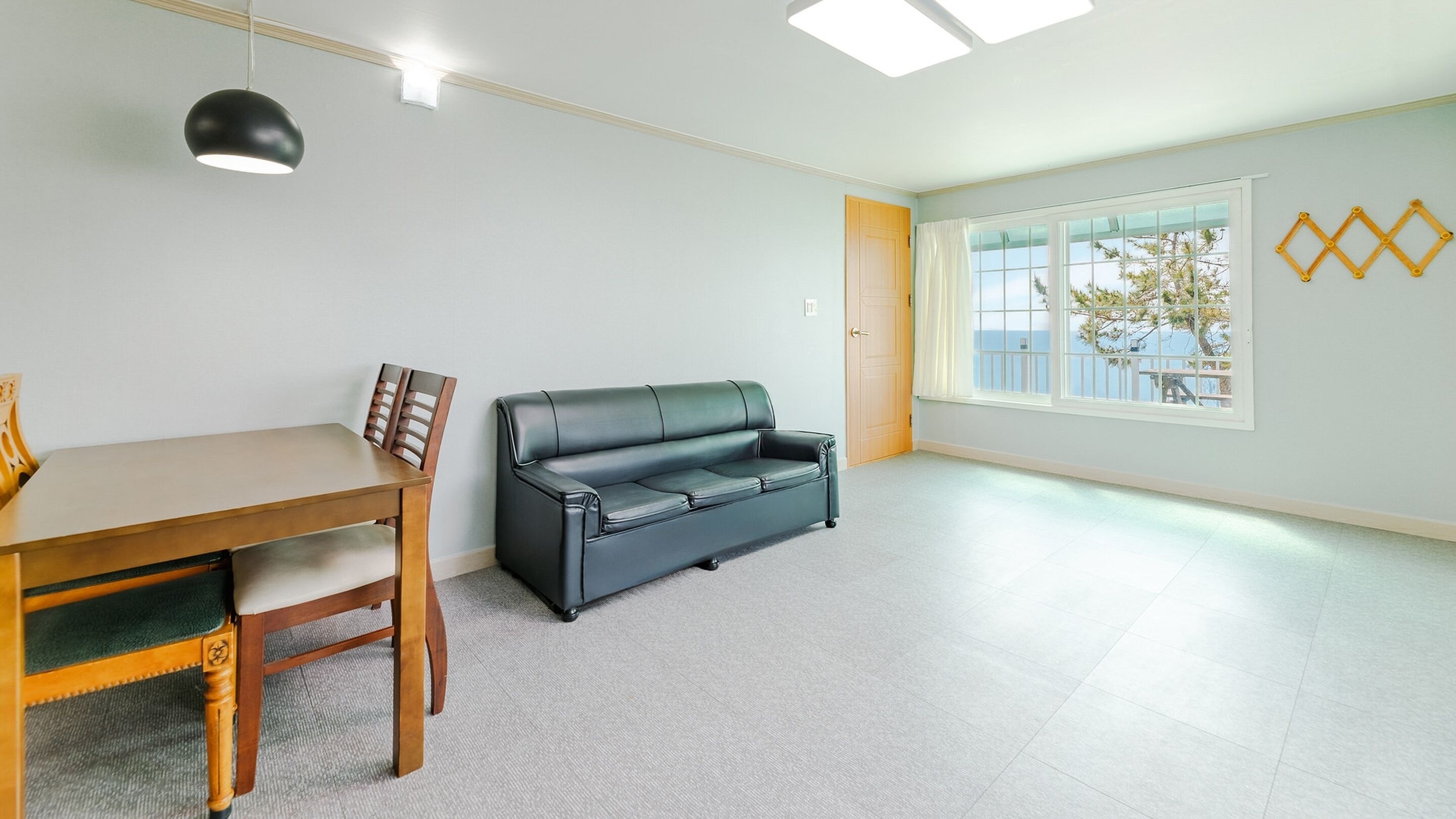 A room with a dining table and chairs, a leather sofa, a large window with a view, and a ceiling light fixture.