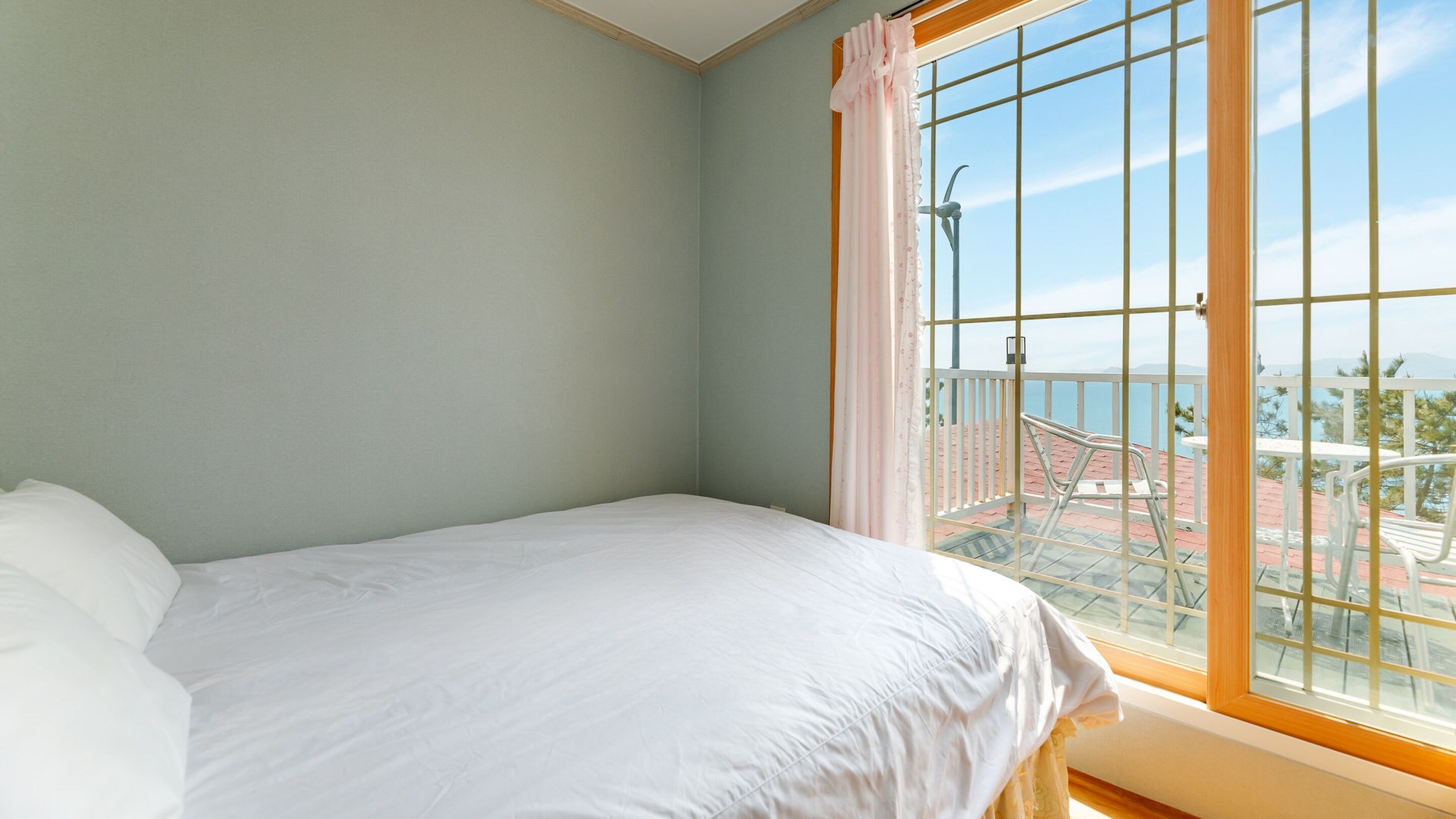 A bedroom with a bed, a window with a view of a beach and wind turbines, and a balcony with a railing.