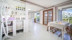 Ruime kamer met een groot raam, een eethoek met een tafel en een witte kast met diverse decoratieve objecten.