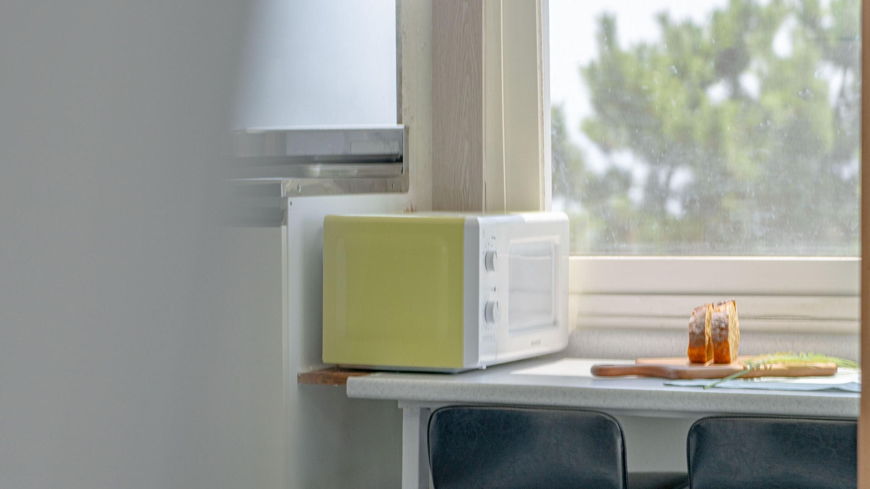 Una cocina con un microondas sobre una encimera, un ventanal con vistas a árboles, y un alféizar con un vaso de jugo de naranja.
