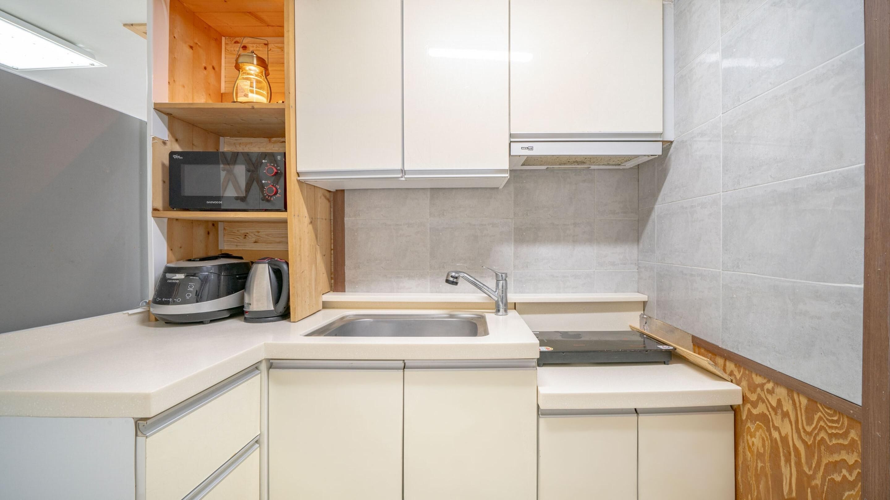 Cocina compacta con armarios blancos, un lavamanos de acero inoxidable, un microondas y una tetera.