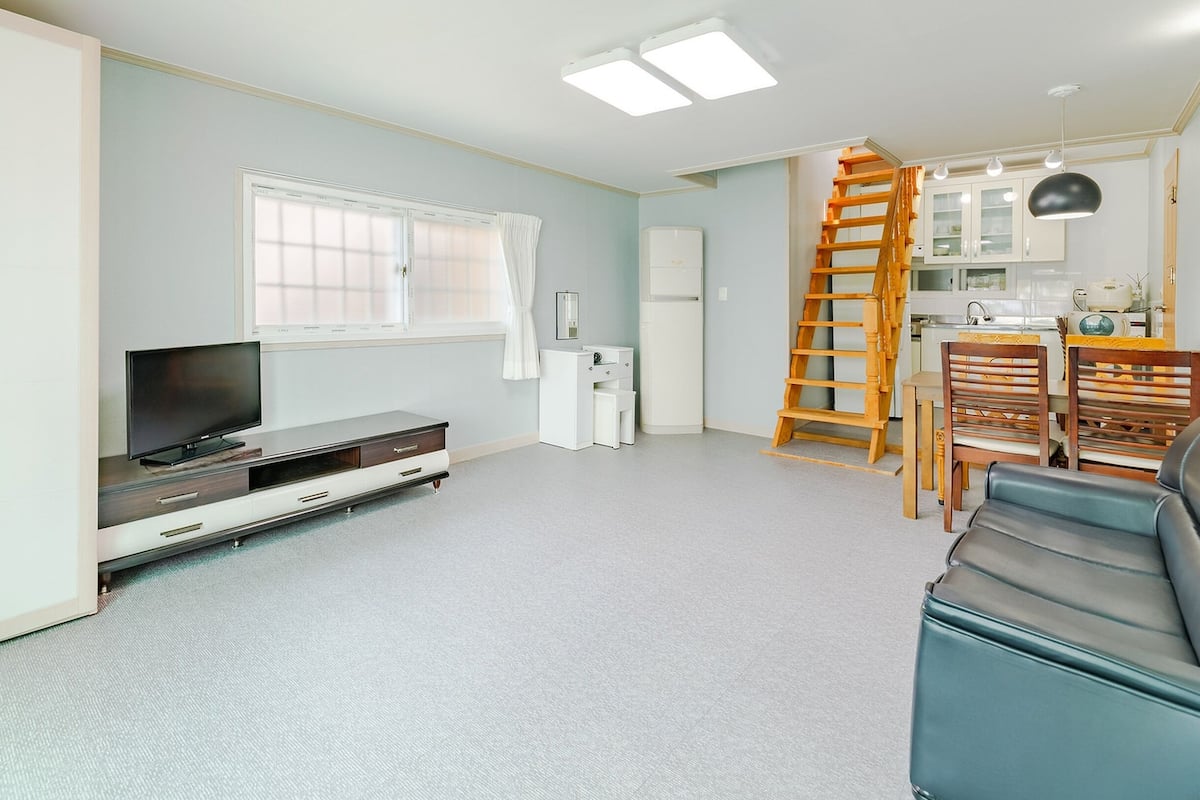 A modern living room with a flat-screen TV, a dining area with wooden chairs, and a kitchen with cabinets and a ladder.