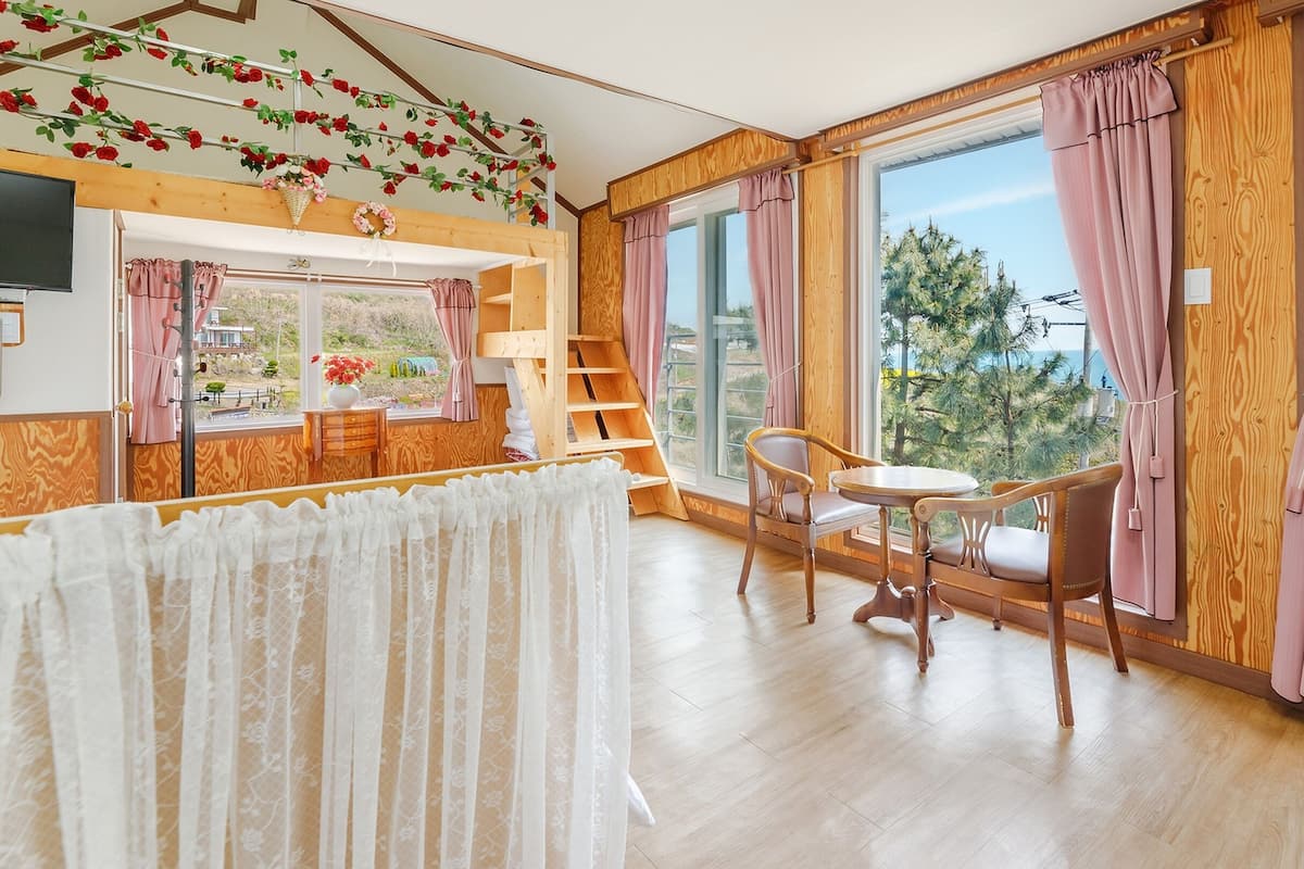 A room with a wooden floor, a window with pink curtains, a table with chairs, and a view of trees outside.
