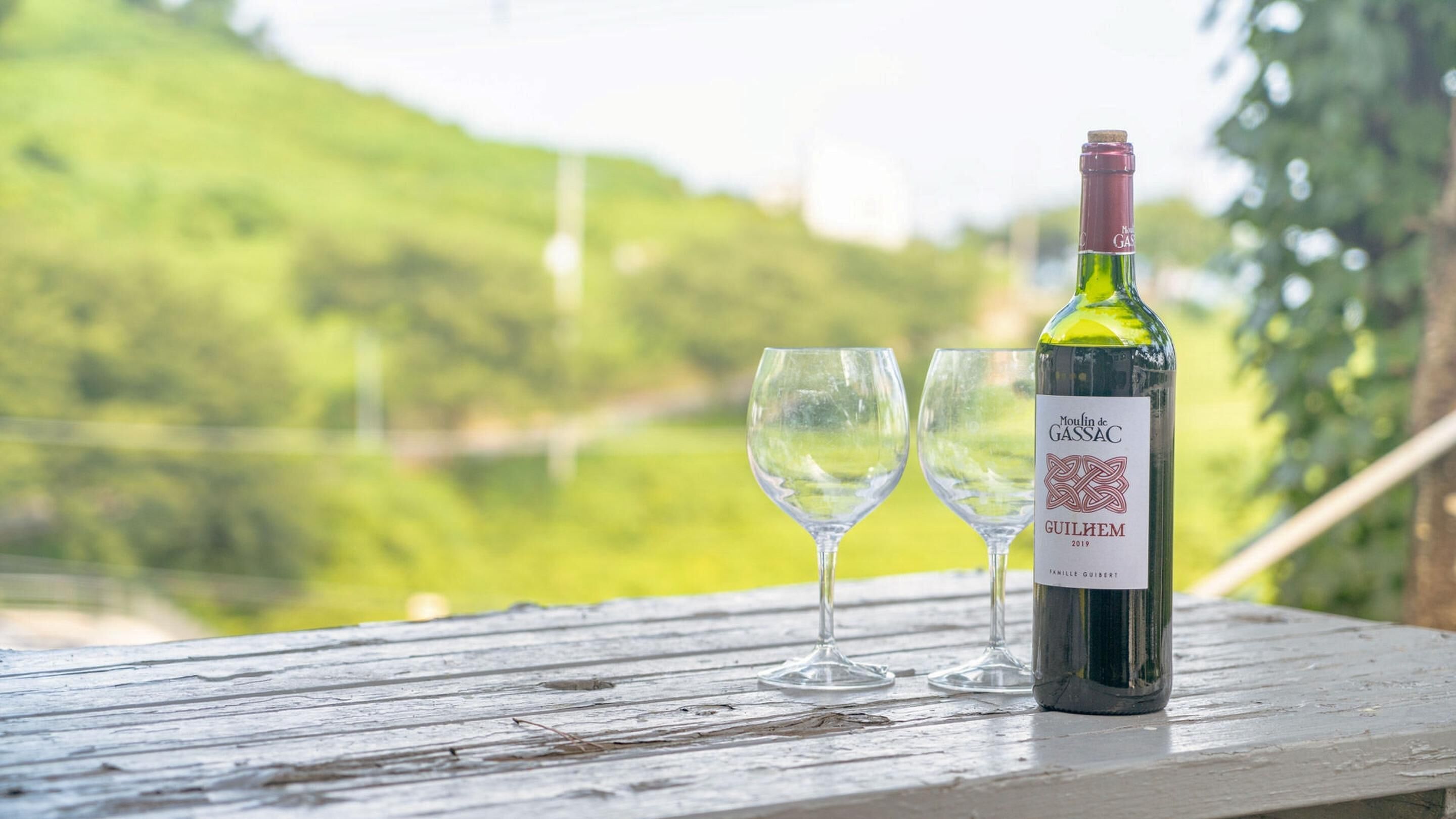 A bottle of Moulin de Gassac GUILHEM 2019 wine on a wooden table with two empty wine glasses, set against a blurred green landscape.