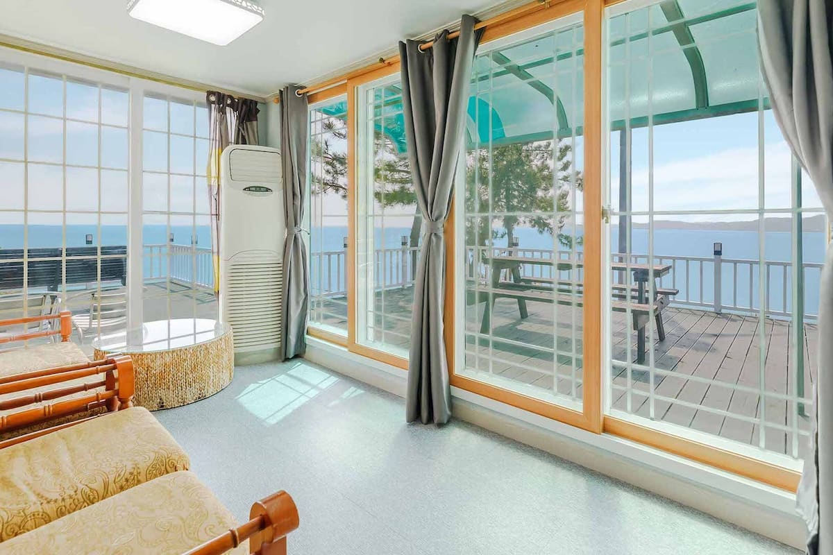 A room with a large window overlooking a deck with a view of the sea. There is a bed with a headboard, a chair, and a small table.
