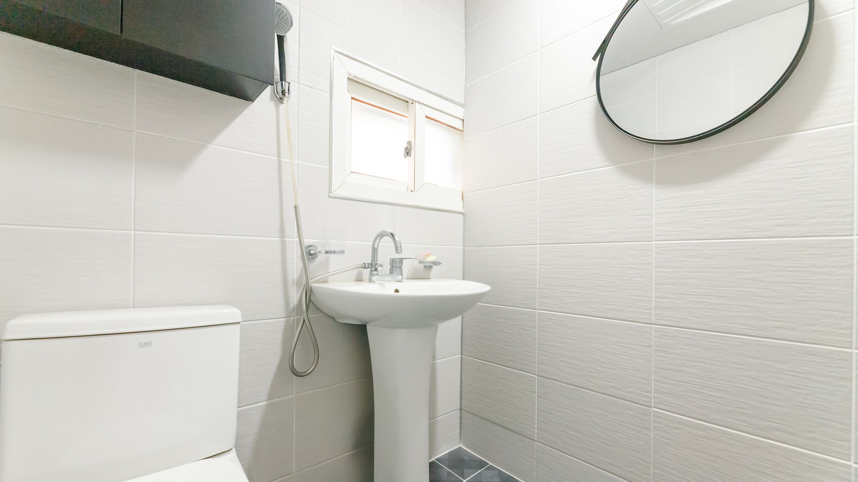 Ein kleines, modernes Badezimmer mit weißen Fliesen, einer Toilette, einem Waschbecken, einer Dusche und einem runden Spiegel.