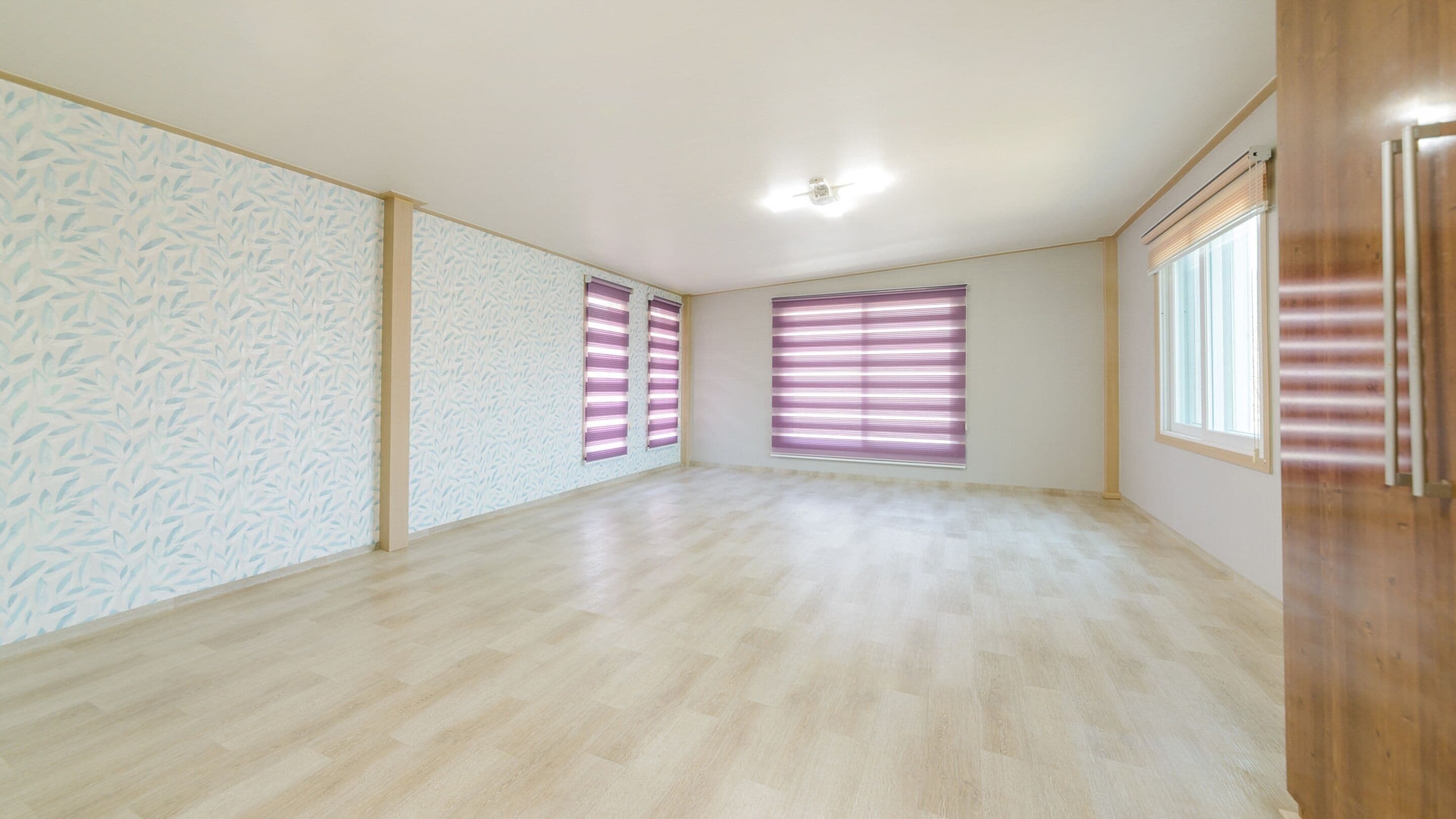 Ein geräumiges Zimmer mit hellem Holzboden, großen Fenstern mit horizontalen Jalousien und einer Deckenleuchte.