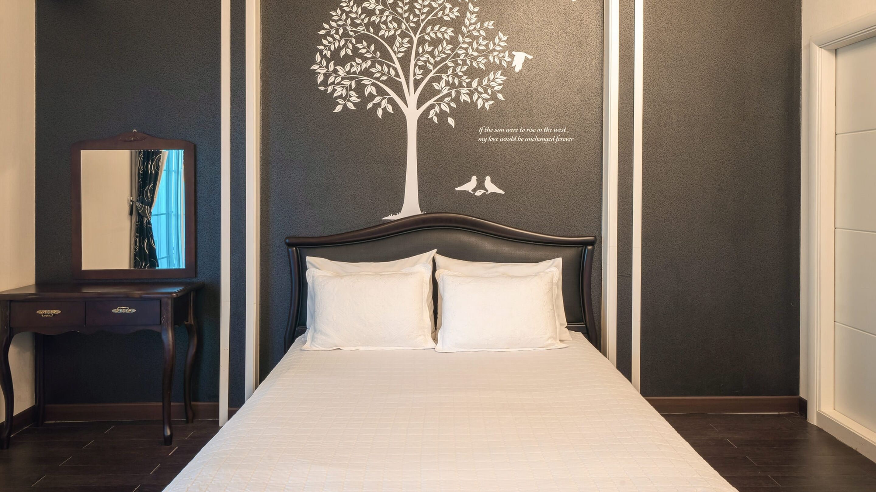 Cama bem arrumada com lençóis brancos em quarto com paredes escuras e um grande adesivo de árvore na parede.