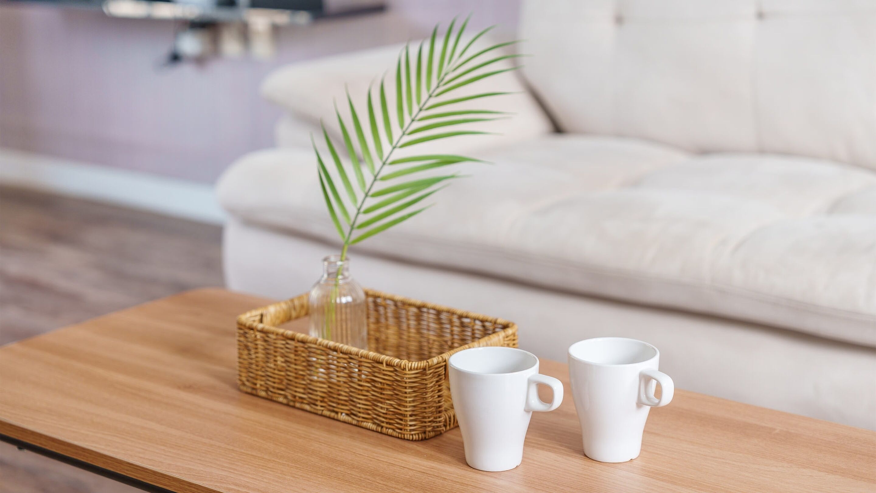 A wooden coffee table with a wicker basket containing a glass vase with a single green plant, and two white ceramic mugs.