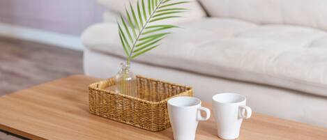 A wooden coffee table with a wicker basket containing a glass vase with a single green plant, and two white ceramic mugs.