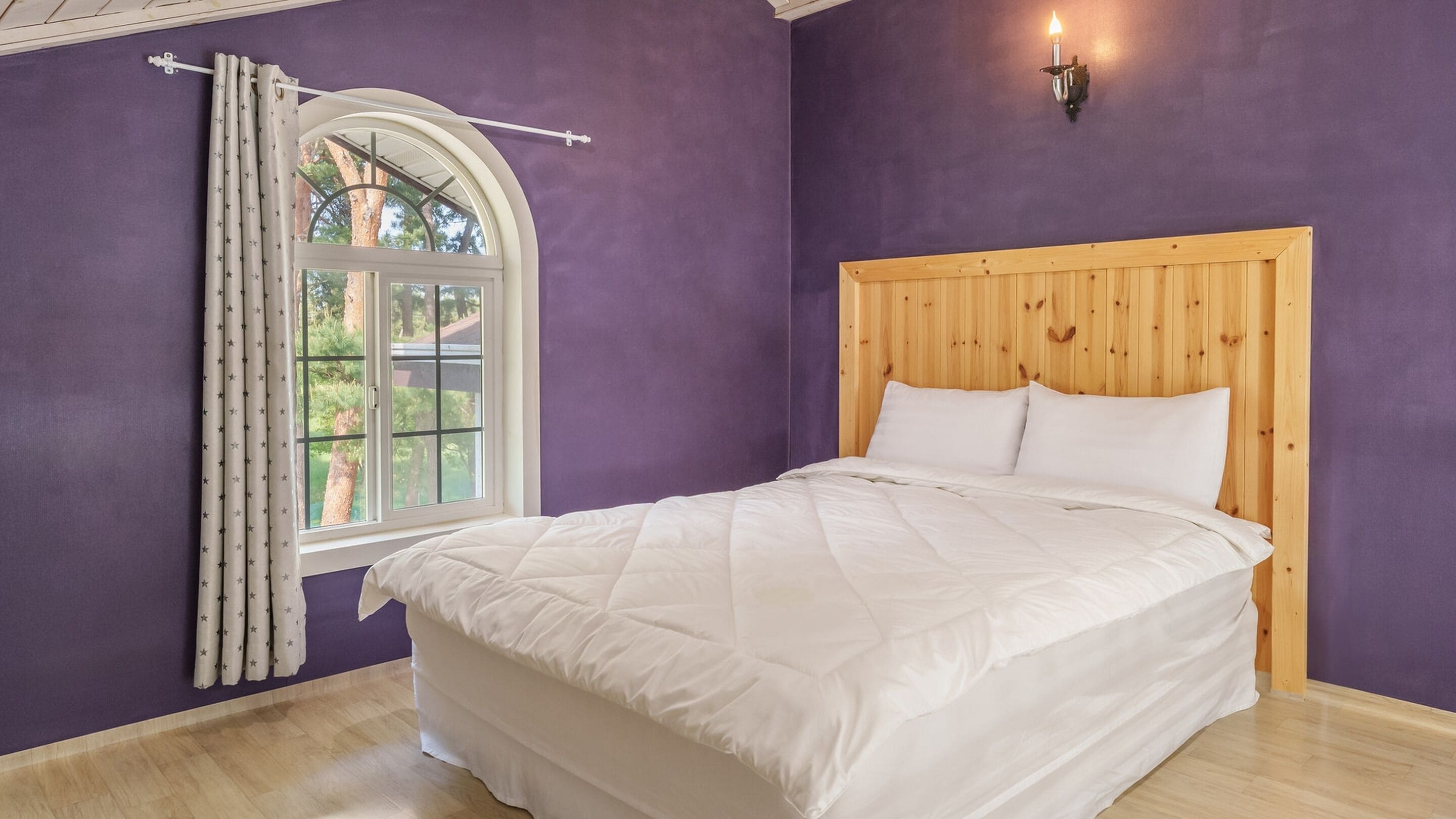 Une chambre à coucher avec une tête de lit en bois, une grande fenêtre avec des rideaux et un lit recouvert de linge de lit blanc.