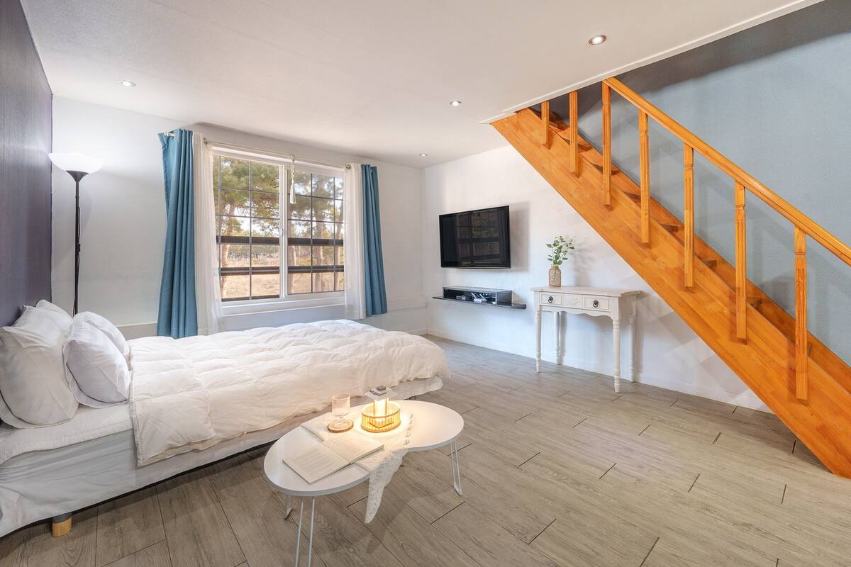A bedroom with a bed, a bedside table, a lamp, a TV mounted on the wall, and a wooden staircase.