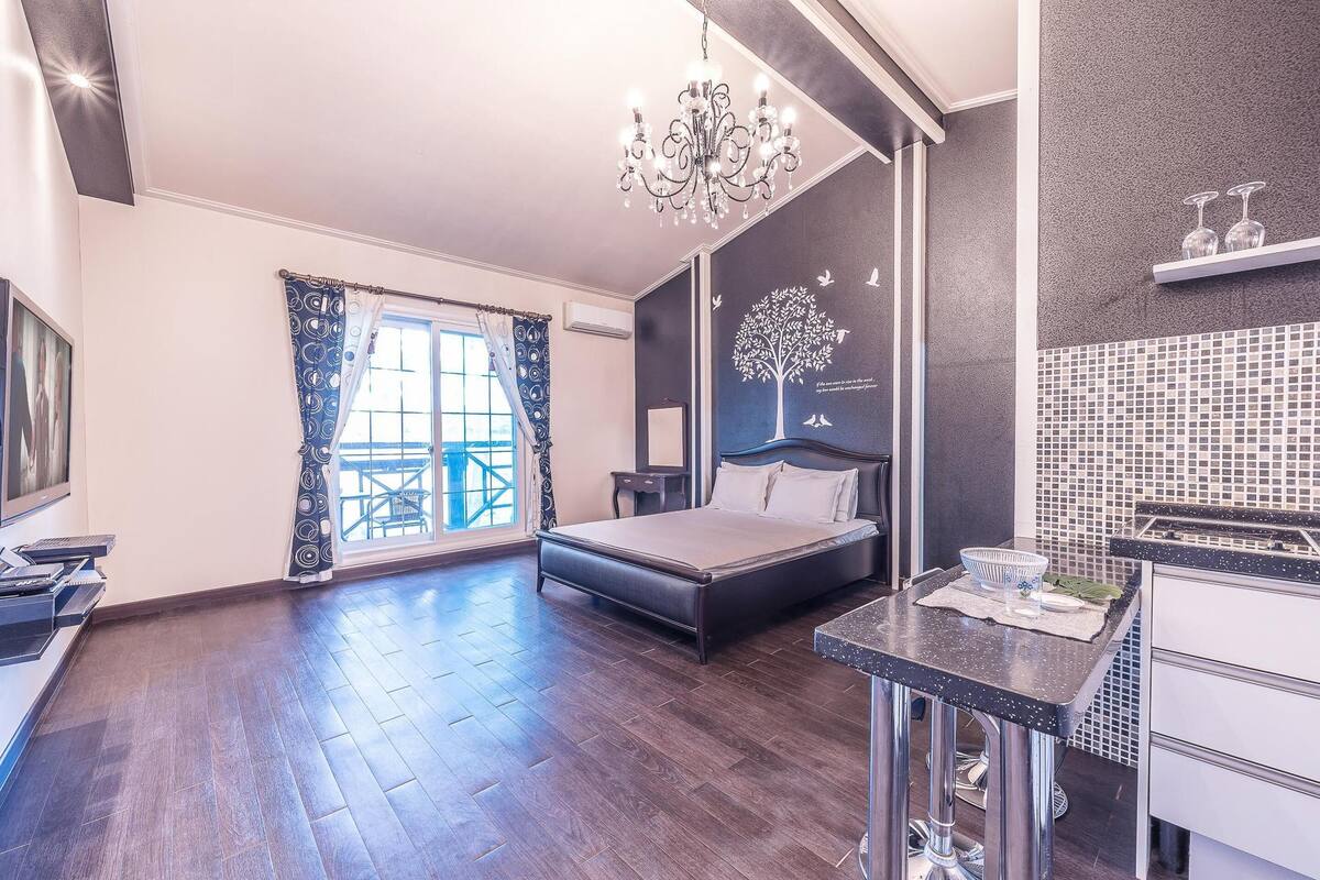 A modern bedroom with a large bed, a chandelier, a view of the outdoors, and a kitchen area with a sink and countertop.