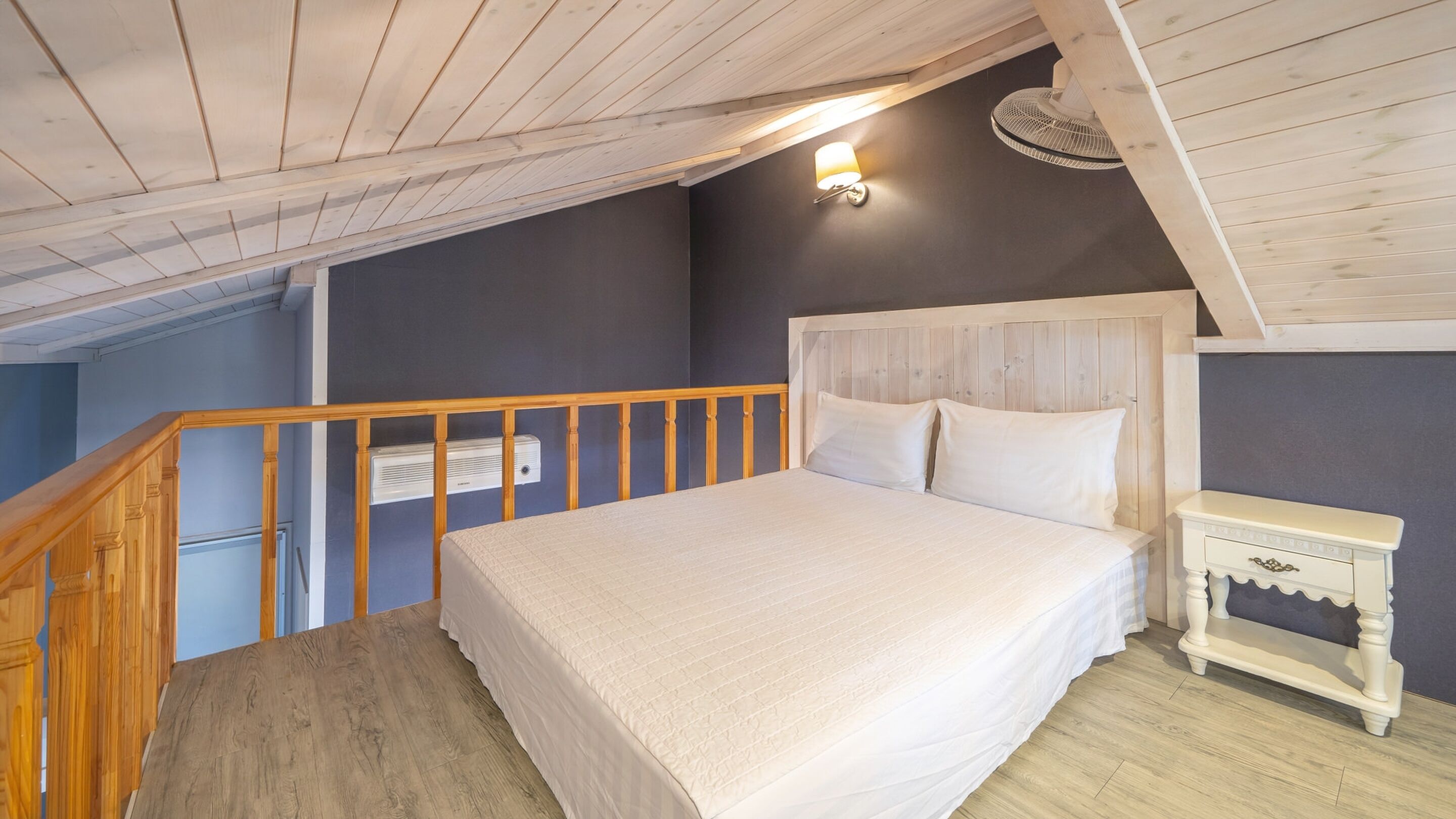 A bedroom with a wooden ceiling, a bed with white bedding, a bedside table, and a wall-mounted light fixture.