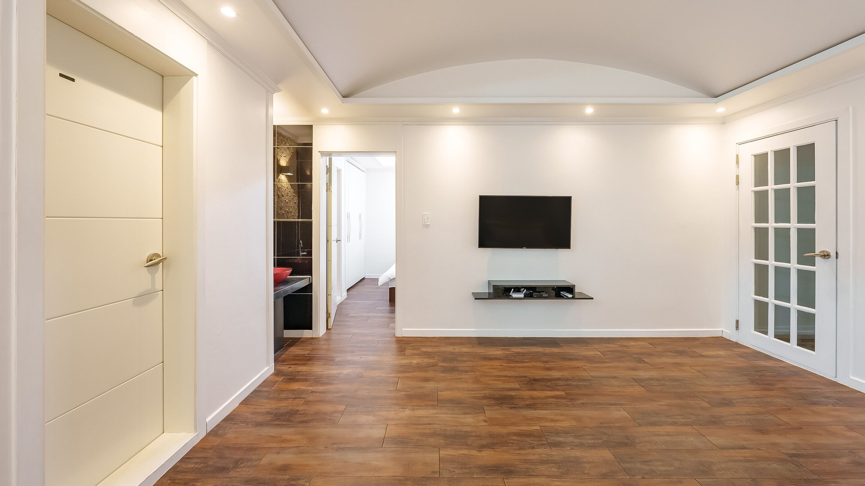 A hallway with a flat-screen TV mounted on the wall, wooden flooring, and a white door with glass panels.