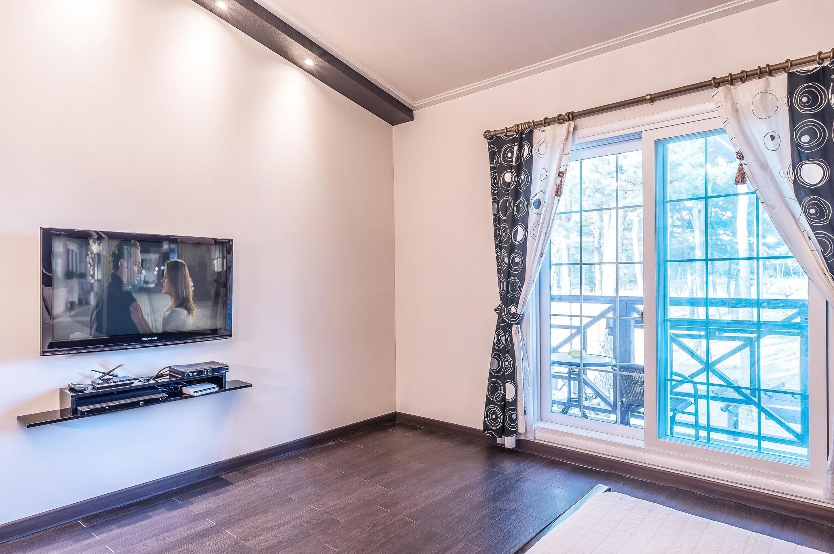 A modern living room with a flat-screen TV mounted on the wall, a built-in entertainment unit, and a large window with patterned curtains.