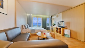 Una habitación de hotel moderna con un sofá, una pequeña mesa redonda con una bebida, un televisor y un balcón con vista al mar.