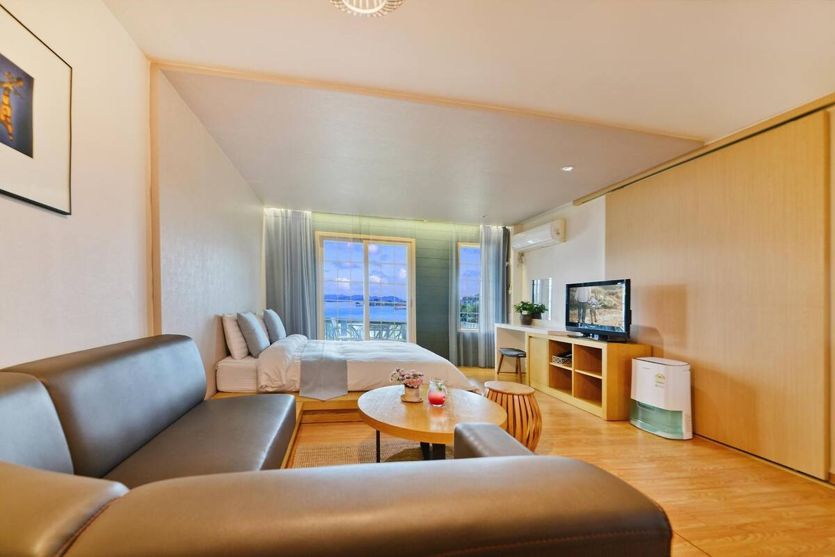 Una habitación de hotel moderna con un sofá, una pequeña mesa redonda con una bebida, un televisor y un balcón con vista al mar.