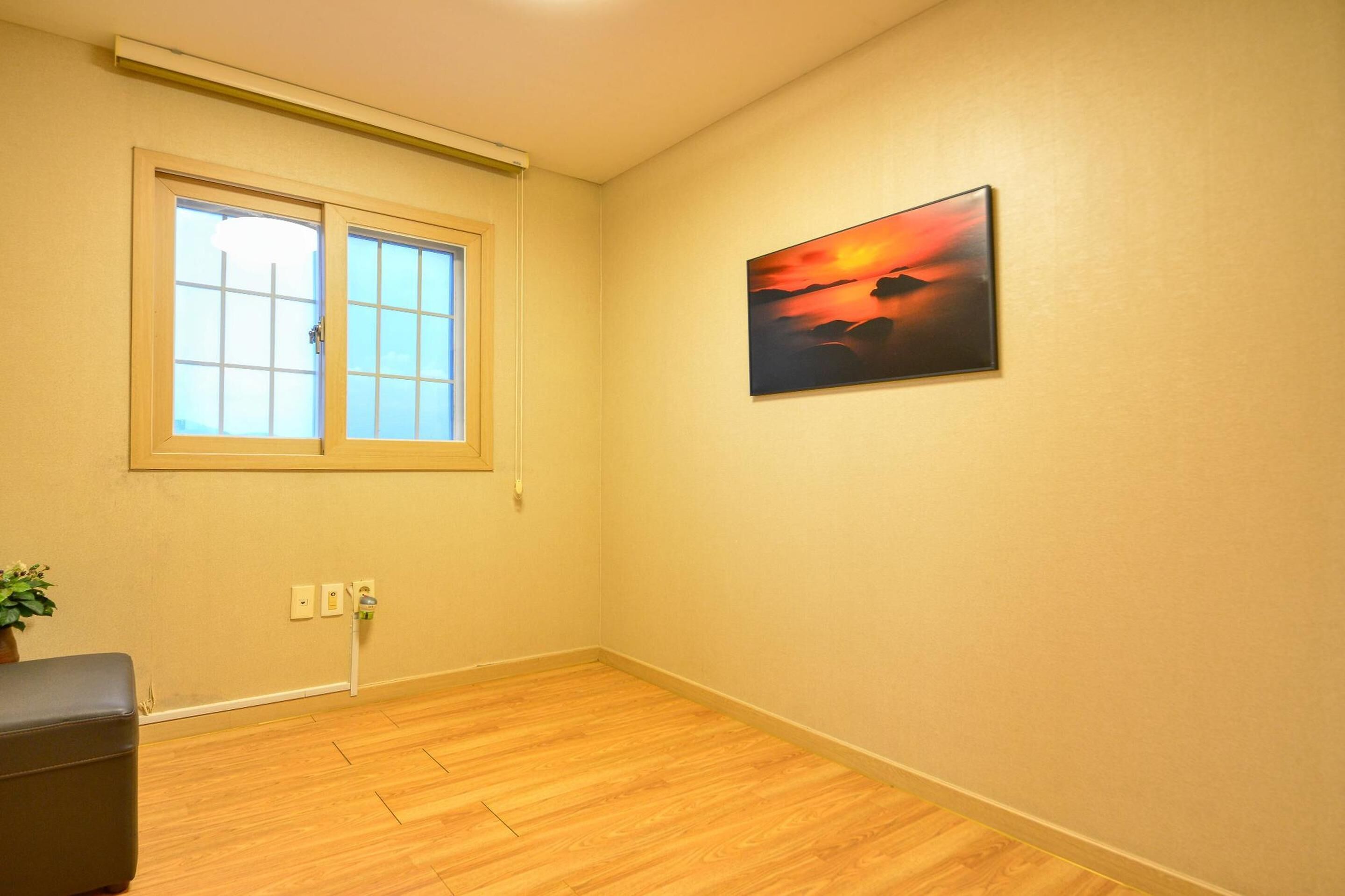 A room with a wooden floor, a window with blinds, a painting on the wall, and a power outlet.