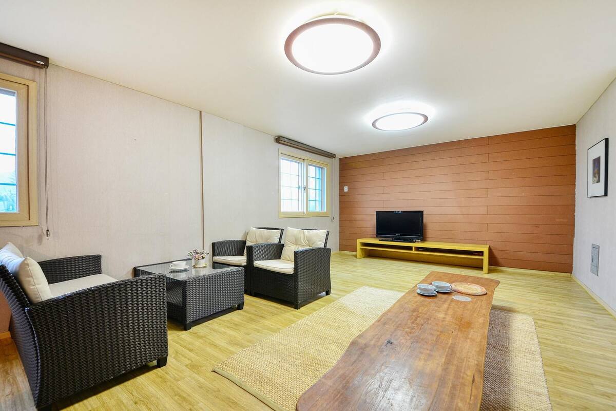 Una sala de estar con piso de madera, dos sofás de mimbre negro con cojines color beige, una mesa de centro de madera, un televisor sobre un mueble de madera y un ventanal con persianas.