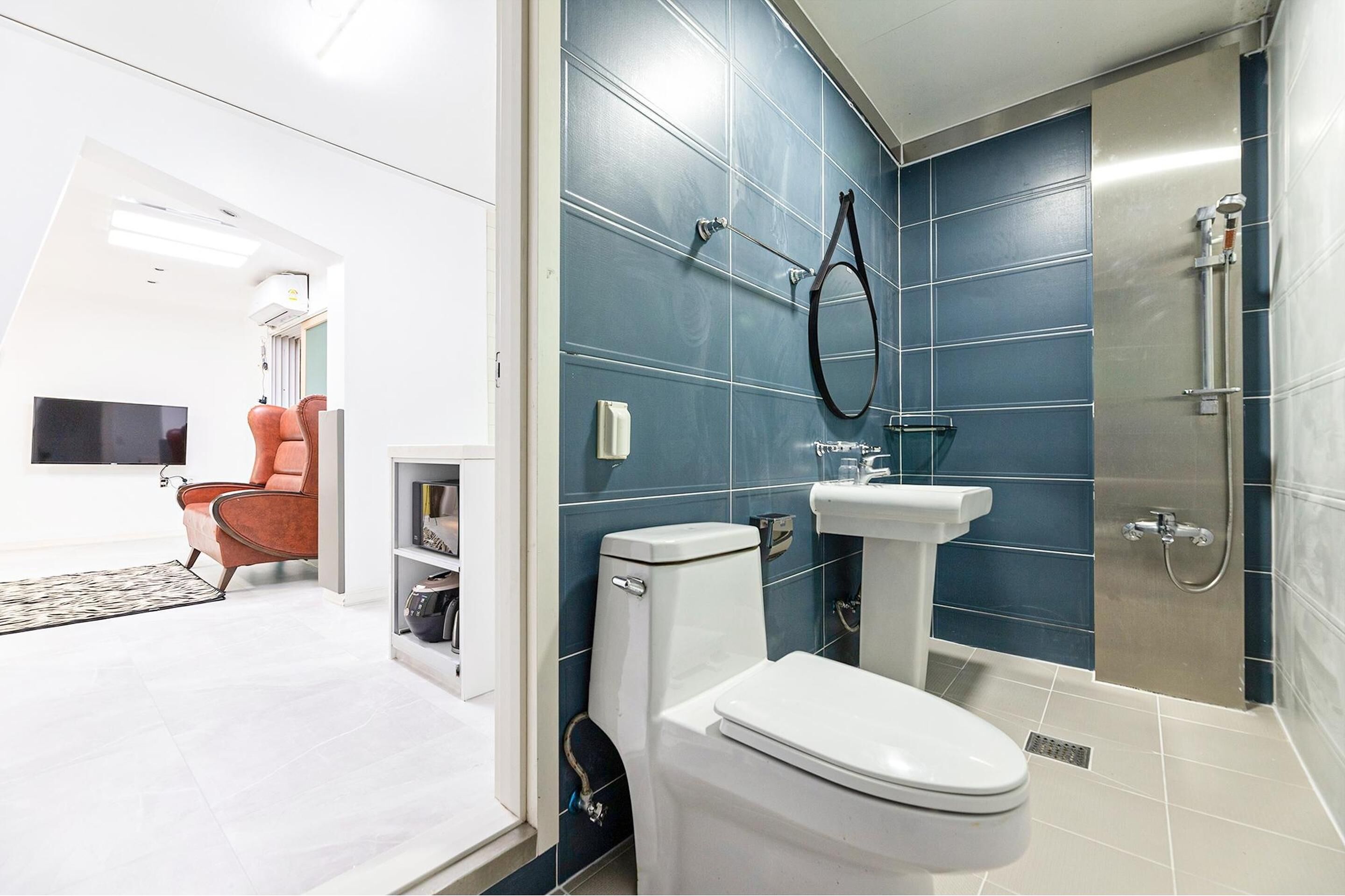 Ein modernes Badezimmer mit Toilette, Waschbecken und Dusche. Im angrenzenden Wohnbereich befindet sich ein Fernseher und ein roter Sessel.