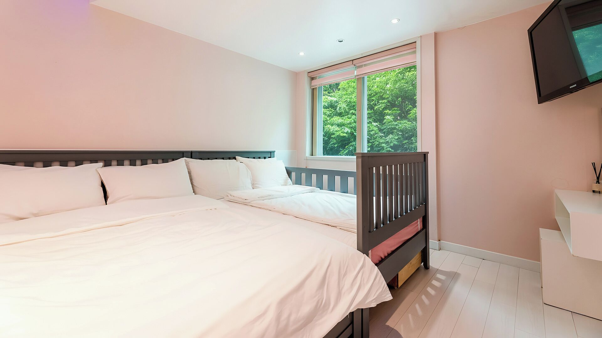 Un dormitorio con cama, ventana con vista a la vegetación, televisor montado en la pared y una pequeña mesa de trabajo con lámpara.