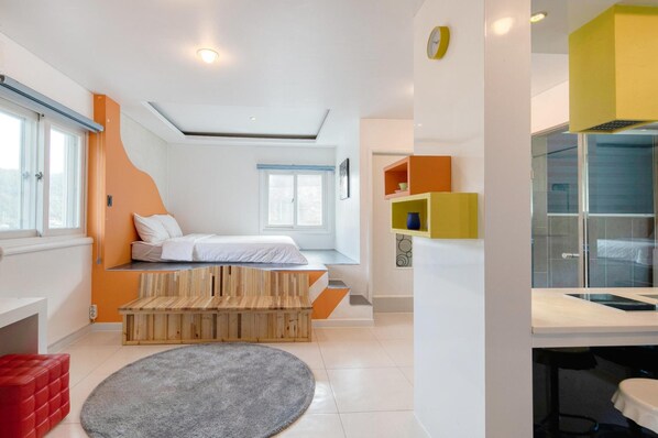 Un dormitorio moderno con una cama, un banco de madera, un reposapiés rojo, una alfombra gris y un baño con cabina de ducha de vidrio.